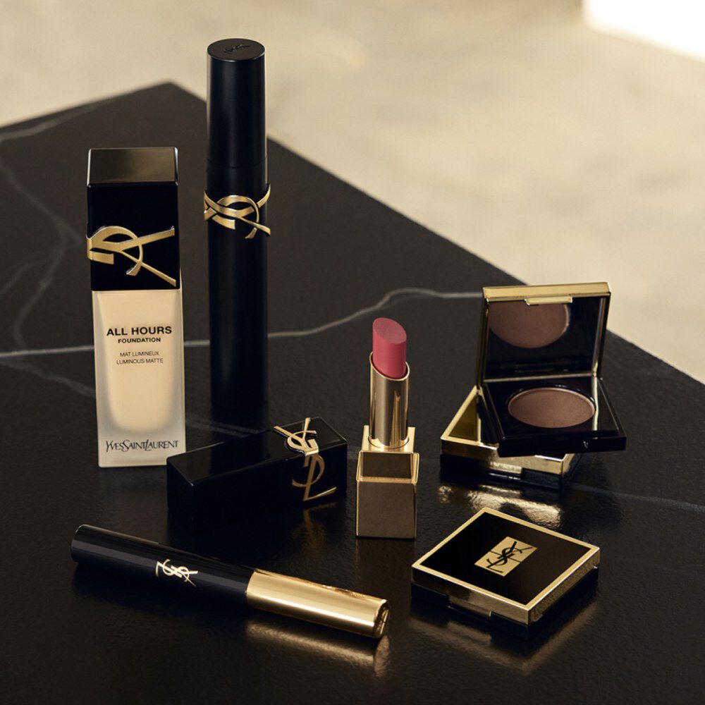 YSL makeup products including foundation, mascara, lipstick, and eyeshadow on a black surface.