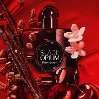 A bottle of Yves Saint Laurent Black Opium perfume surrounded by coffee beans, vanilla pods, and flowers on a red background.