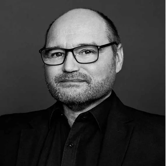 A person in a black suit against a dark background.