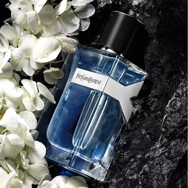 A rectangular YSL perfume bottle with blue liquid surrounded by white flowers on a black marble background.