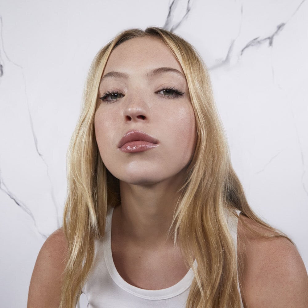 A person with long blonde hair wearing a white tank top against a marble background.