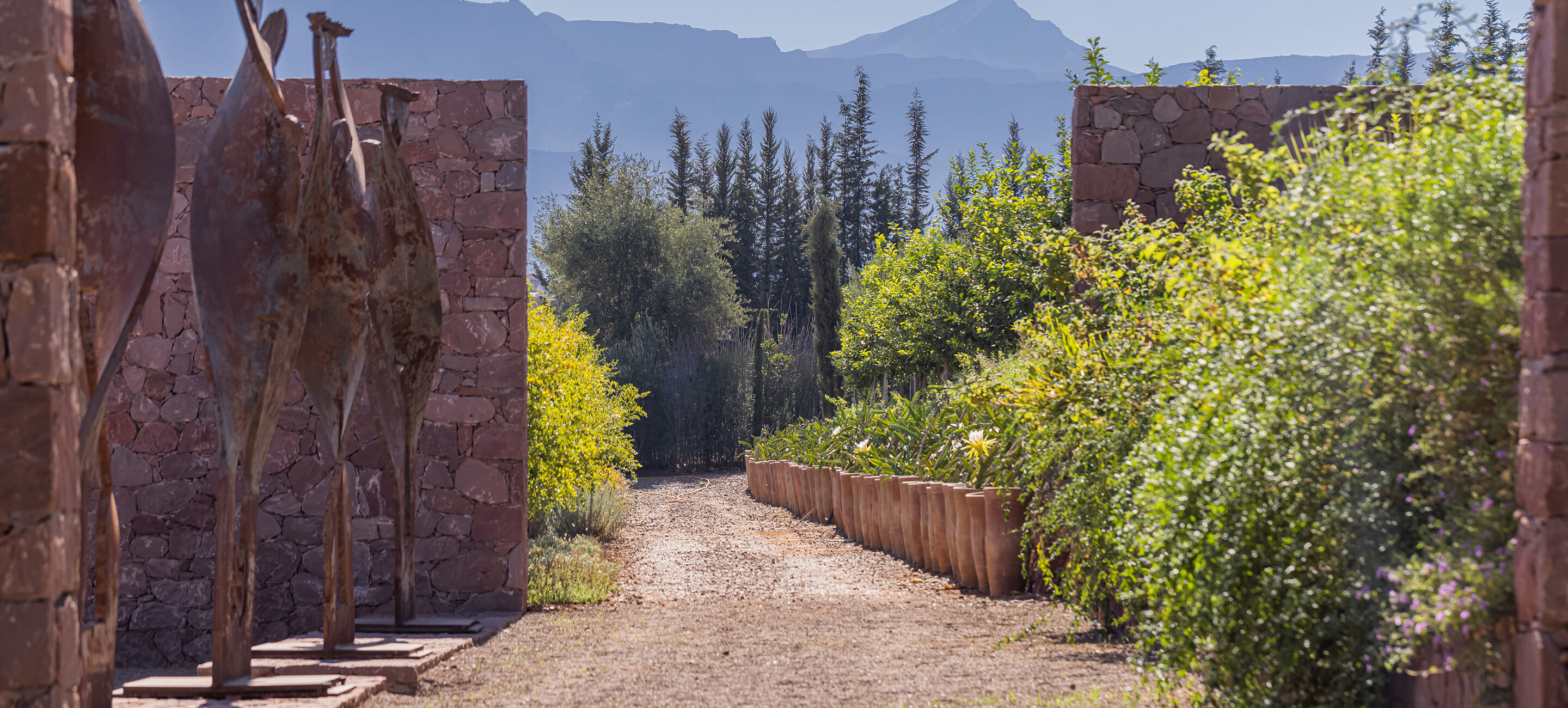 Pathway lined with plants and sculptures leading to mountains in the distance.