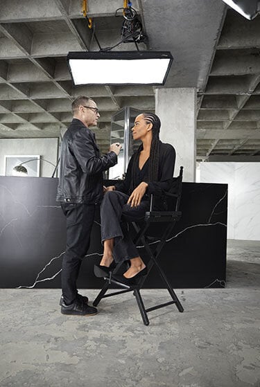 Makeup artist applying makeup to a person sitting on a high chair in a studio.
