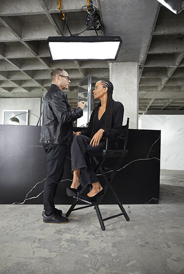 Makeup artist applying makeup to a person sitting on a high chair in a studio.