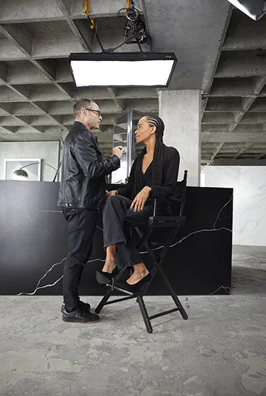 Makeup artist applying makeup to a person sitting on a high chair in a studio.