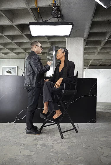 Makeup artist applying makeup to a person sitting on a high chair in a studio.
