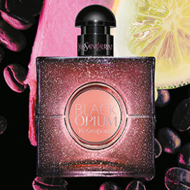 A bottle of YSL Black Opium perfume with a pink center, set against a background of coffee beans and a slice of lime.