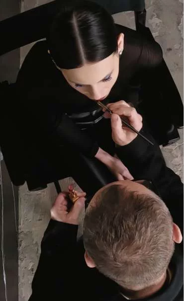 A person applying makeup to another person seated in a chair.