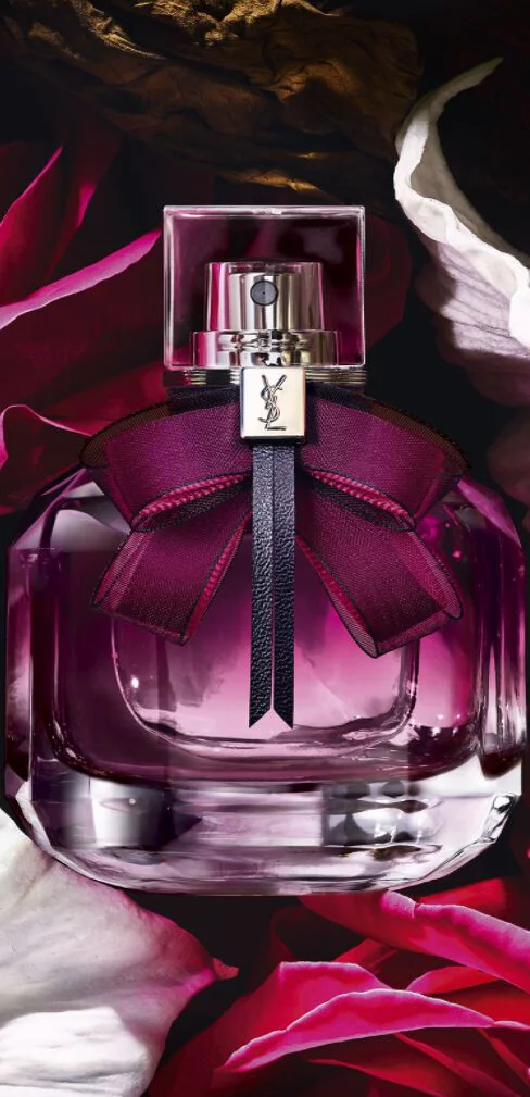 Rectangular YSL perfume bottle with dark purple liquid and a purple bow, surrounded by flowers.