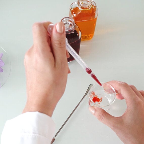 A person using a pipette to add red liquid to a small glass container, with two bottles of amber liquid in the background.
