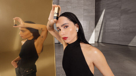 A woman in a black dress holding a YSL product near her face, with her reflection in a gold mirror.