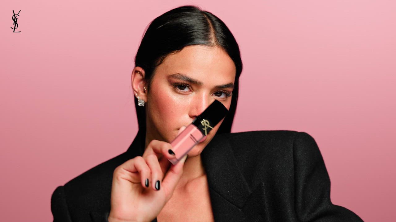 A woman with black hair and earrings in a black outfit against a pink background with a YSL logo in the corner.
