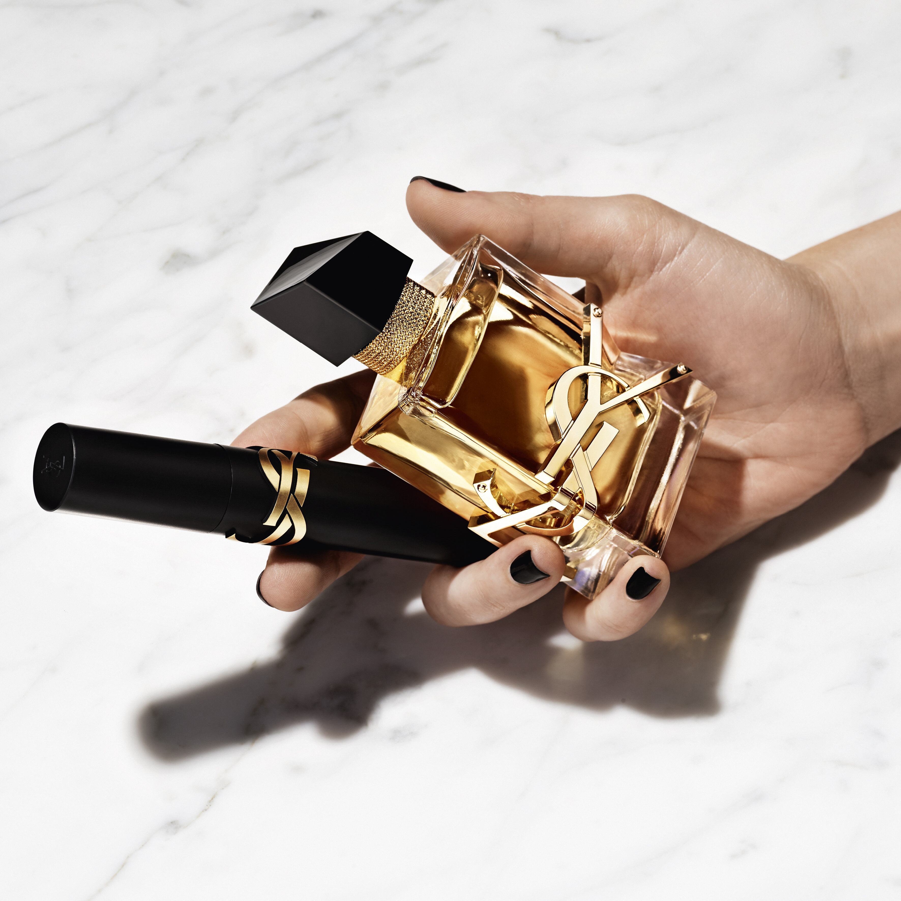 Hand holding a YSL perfume bottle and a black YSL product on a marble surface.