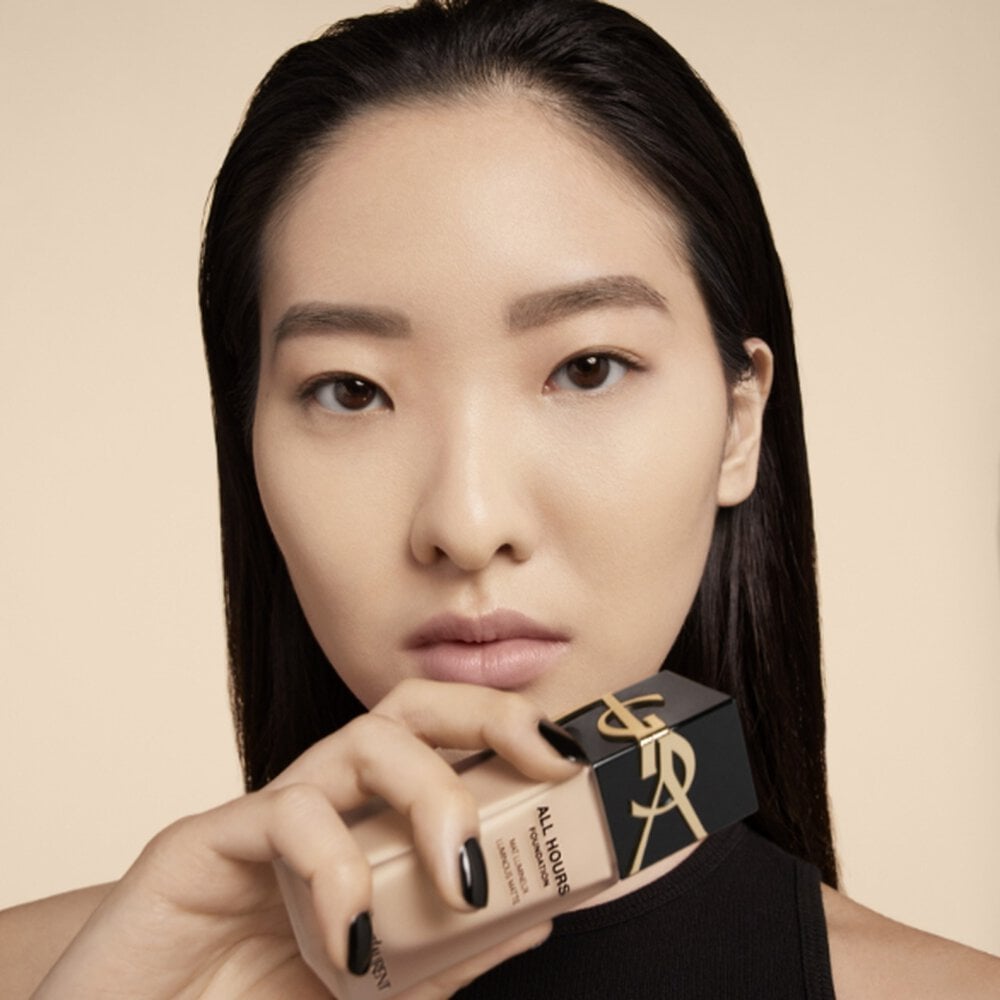 A woman holding a bottle of YSL All Hours foundation near her face.