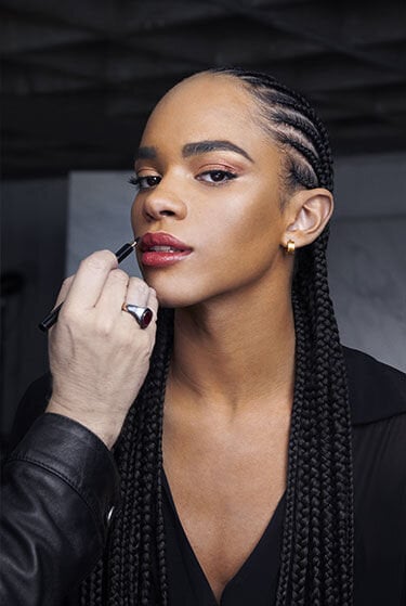 A makeup artist applies makeup to a person with long braided hair.