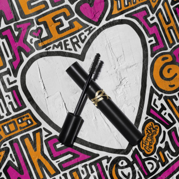 A black tube of mascara with a wand placed on a colorful graffiti-style background featuring a large white heart.