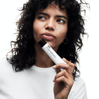 A person with curly hair holds a white Yves Saint Laurent product near their face.