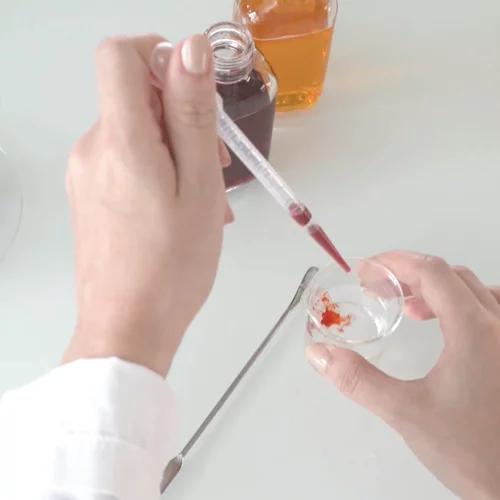 Hands using a dropper to mix liquids in a lab setting with flowers and beakers.