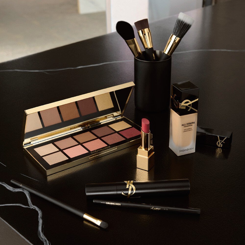 An exquisitely styled YSL-inspired makeup still life arranged on a glossy black surface that reflects every object like a dark mirror. Deep red lipsticks, gold-capped mascara, a black and gold eyeshadow palette with shimmering jewel tones, and an elegant glass bottle of nail lacquer are positioned with geometric precision. A single white gardenia lies beside them, its velvety petals contrasting with the hard metallic lines. Dramatic, directional studio lighting from the side creates high-contrast highlights and inky shadows, with luminous bokeh from out-of-focus city lights in the distant background. Photographic realism, low-angle close-up emphasizing texture and reflection, evoking the seductive, nocturnal glamour of a luxury beauty campaign without any human presence.