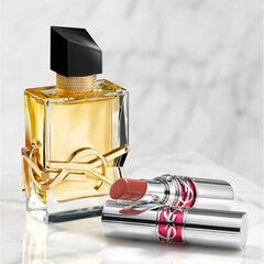 A rectangular YSL perfume bottle with amber liquid and a gold logo sits next to two silver YSL lipsticks on a marble surface.