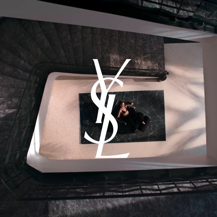 A woman in a black outfit lying on a black marble surface, viewed from above with a large YSL logo overlay.
