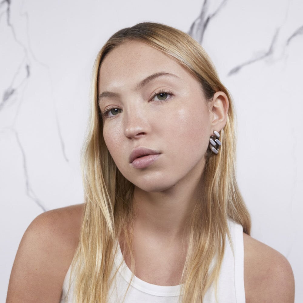 A person with long blonde hair wearing a white tank top and black and white earrings.