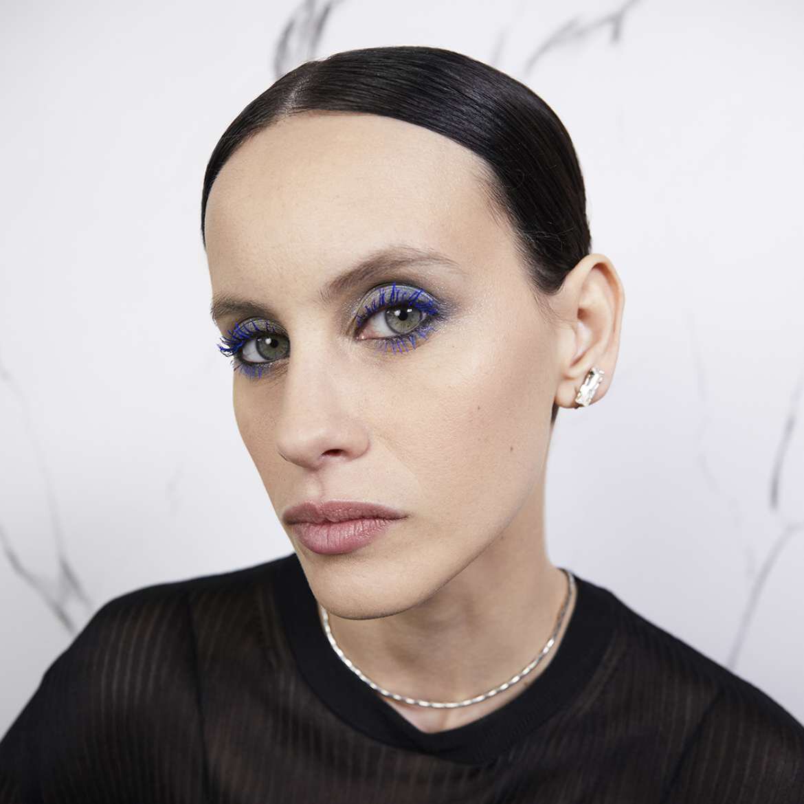 A person with slicked-back hair wearing a black top and silver jewelry.