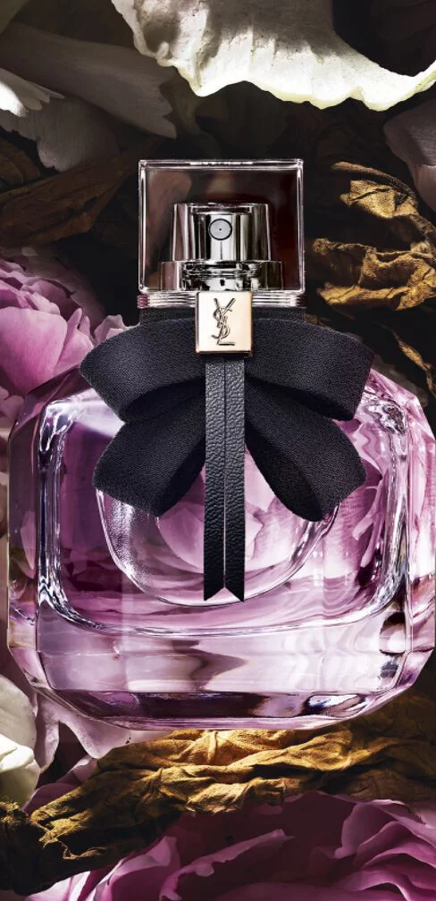 Rectangular YSL perfume bottle with purple liquid and a black bow, surrounded by flowers.