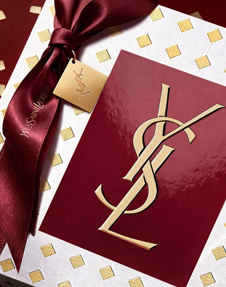 White and gold diamond-patterned YSL gift box tied with a deep burgundy satin ribbon and gold tag, featuring a glossy red center panel with the gold YSL logo.