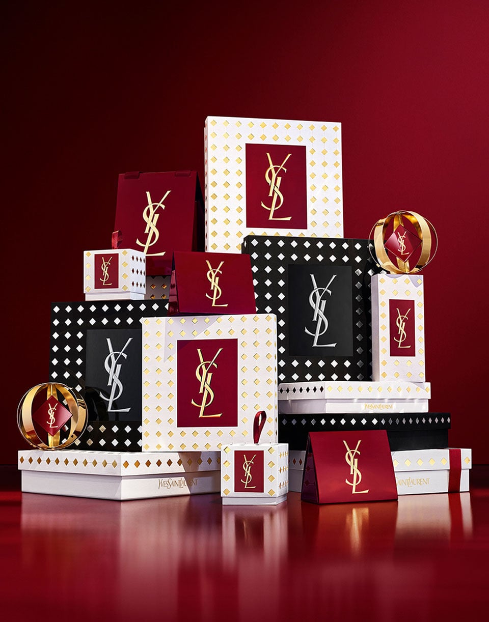 A luxury stack of YSL holiday gift boxes in white, gold, burgundy, and studded black patterns with metallic logos, arranged on a reflective red surface.