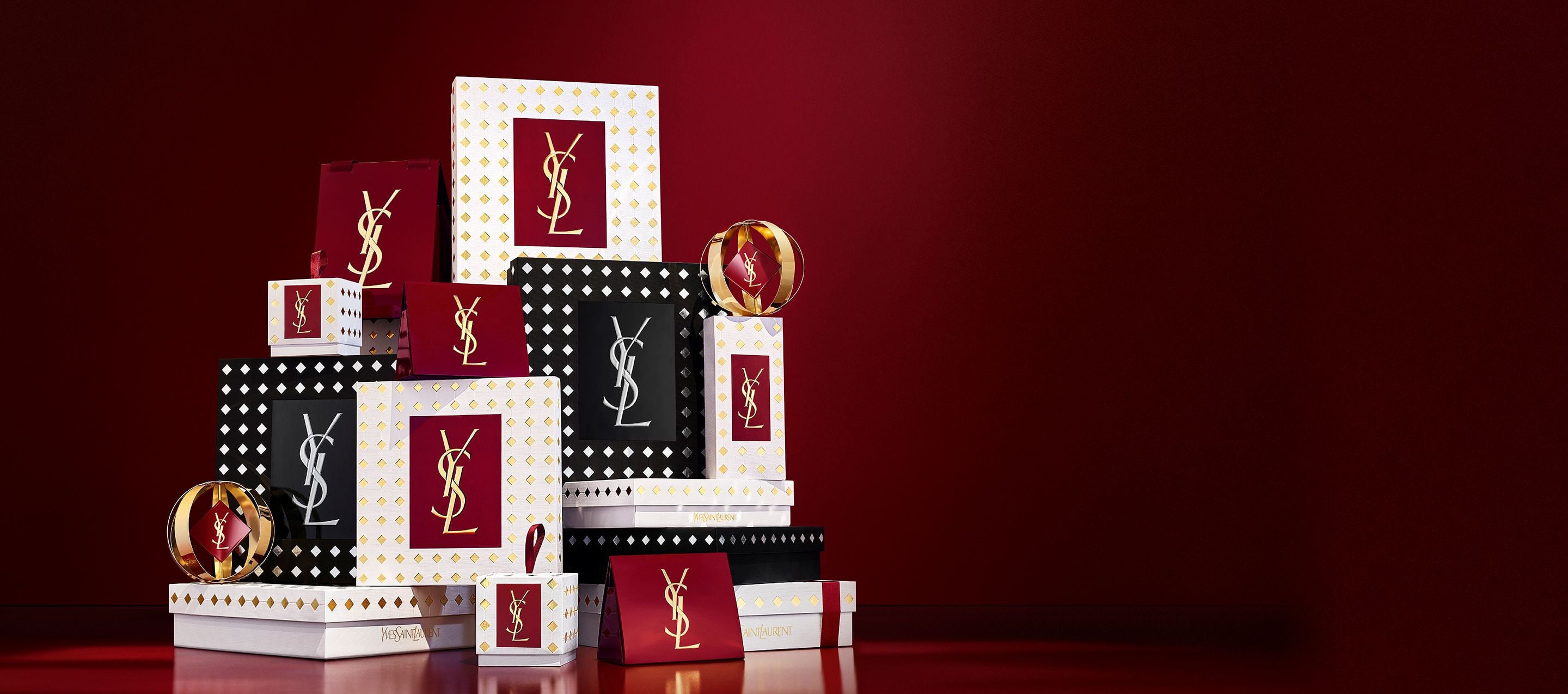 Three models in elegant black outfits holding YSL holiday gift boxes in front of a festive red curtain background with falling gold confetti.